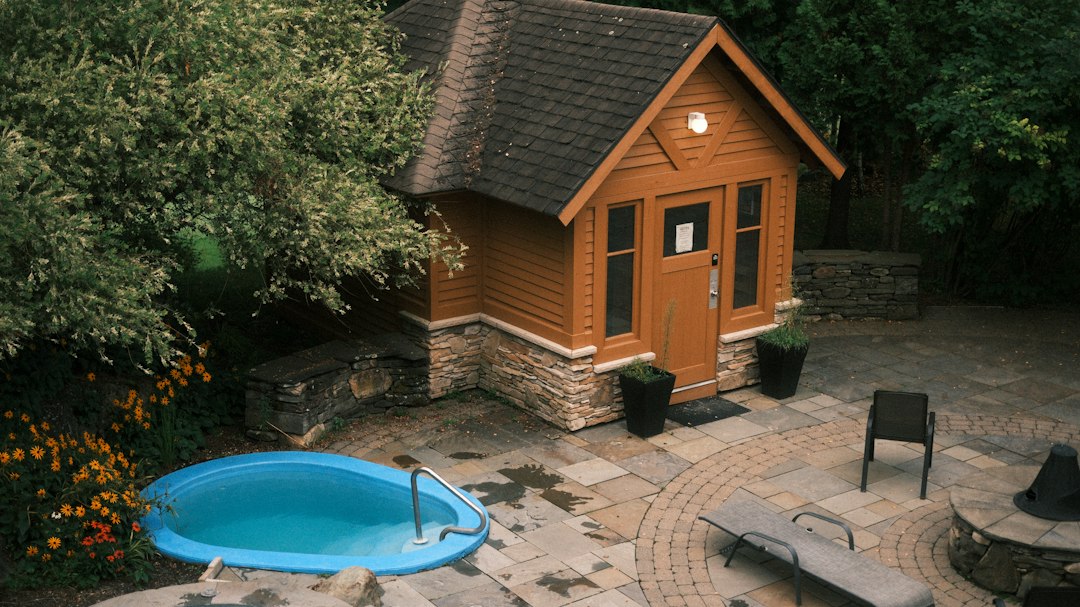 A small wooden cabin with a stone base beside a blue plunge pool on a stone patio